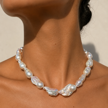 Load image into Gallery viewer, A close-up of a person's neck wearing a baroque pearl necklace. The necklace features irregular, lustrous white pearls with subtle iridescent pearly highlights, set against a warm beige background.

